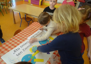 Czwórka dzieci stoi wokół stolika na którym leży plakat ekologiczny, jedna z dziewczynek nakleja hasło, druga elementy z kolorowego papieru.